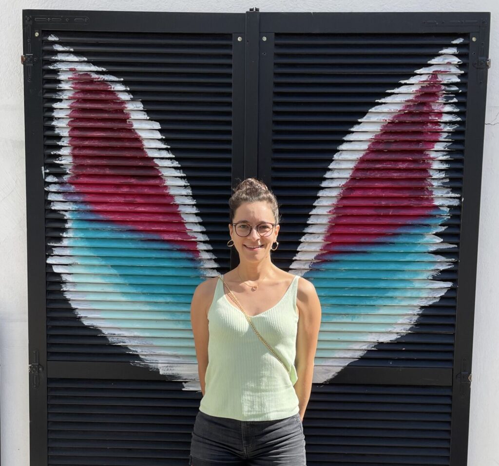 Portrait im Stehen von Barbara Weißensteiner mit bunten Engelsflügeln im Hintergrund an der Wand