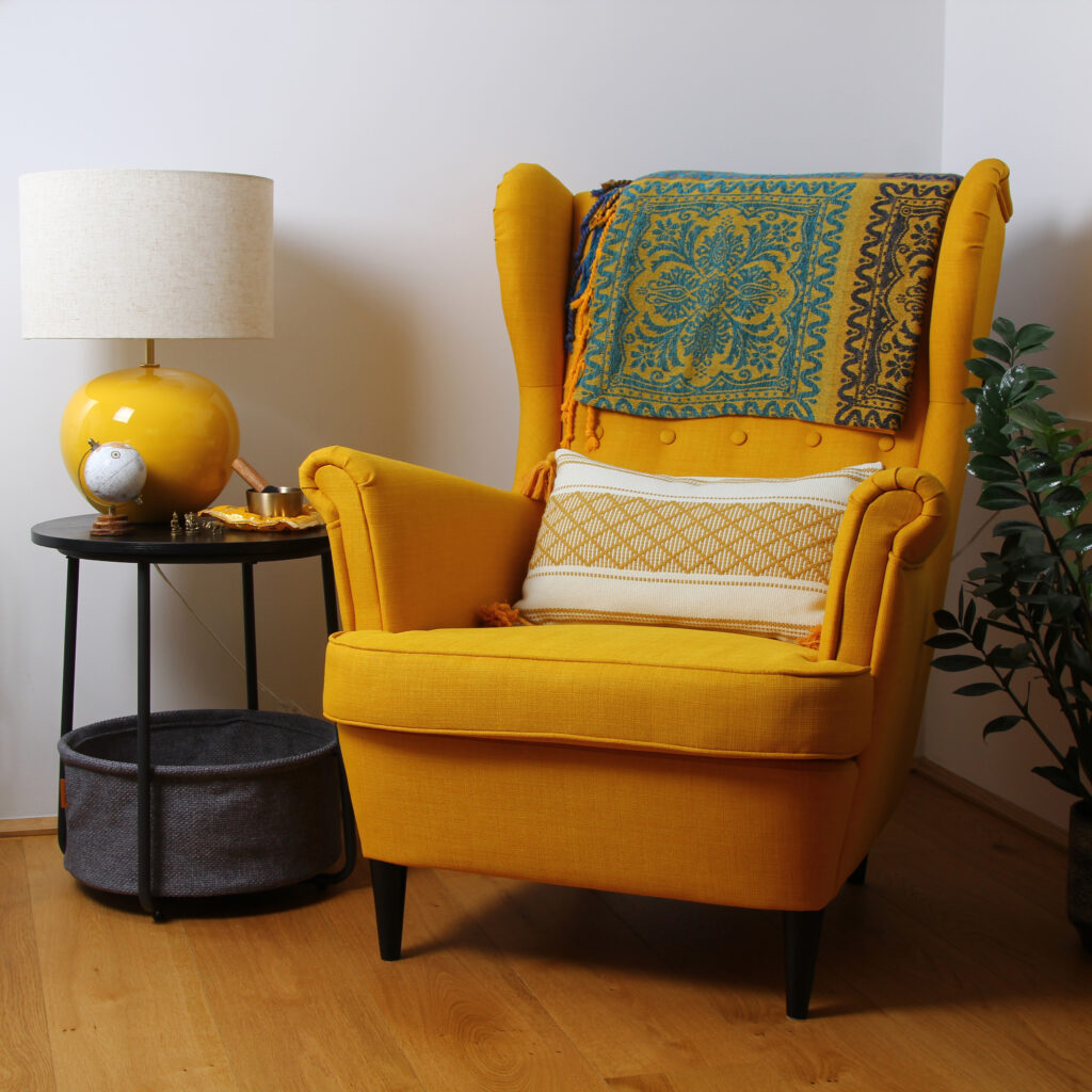 Gelber Ohrensessel mit Polster und Decke daneben schwarzer kleiner Couchtisch mit gelber Lampe