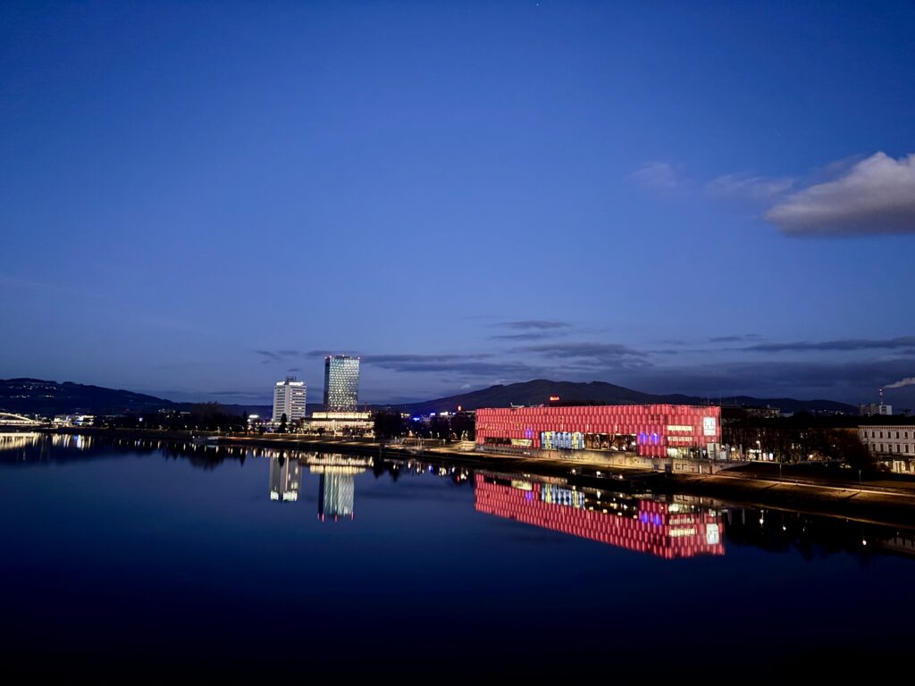 Blick auf die Donau in Linz in der Abenddämmerung, rechts rot leuchtend das Lentos und Hochhäuser