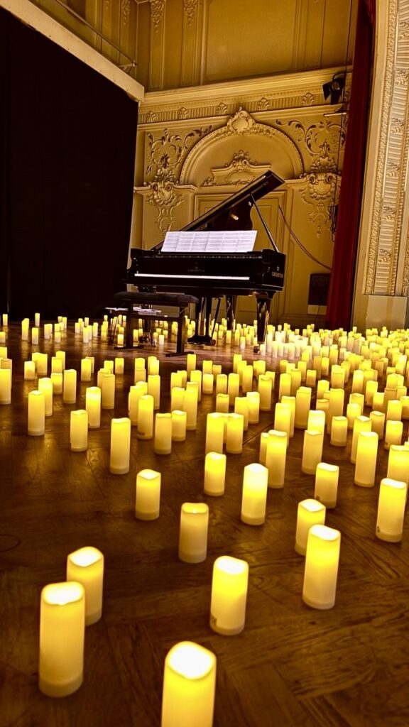 Viele leuchtende Kerzen am Boden verteilt, im Hintergrund ein Klavierflügel mit Noten